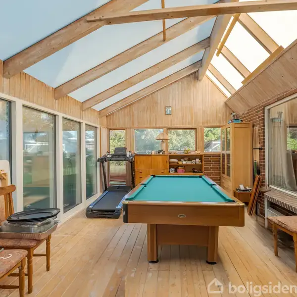 Et billardbord står i midten af en lys udestue med skråt tag. Rundt om er der træmøbler, en løbebånd og store vinduer med udsigt til naturen udenfor.