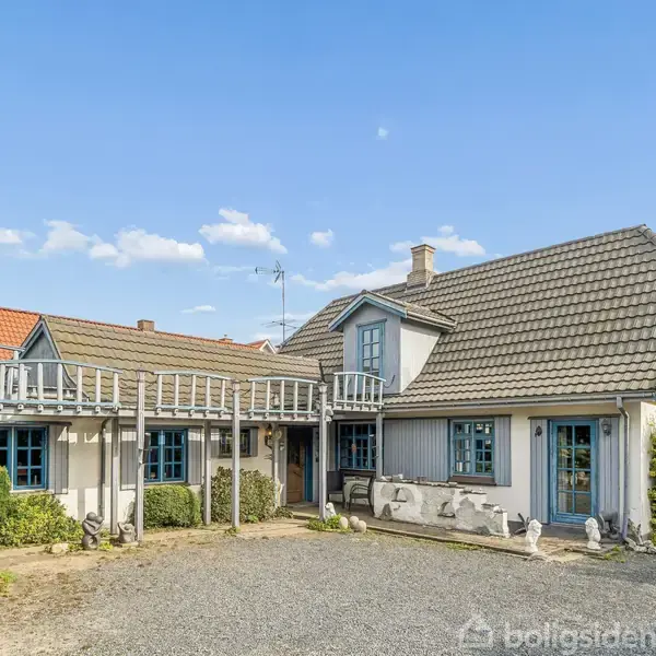 Et traditionelt, grå-hvidt hus med blå vinduer står stille på en grusvej. Omgivet af grønne buske og marker under en klar blå himmel.