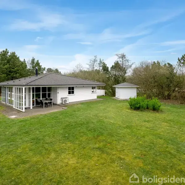 Et hvidt hus med et sort tag står i en græsplæne. Ved siden af huset er et lille hvidt skur. Omkring er der buske og træer under en blå himmel.
