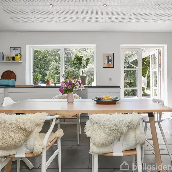 Et lyst køkken med et stort bord omgivet af stole med bløde skind. Friske blomster er i en vase på bordet. Store vinduer og en åben dør fører ud til haven.