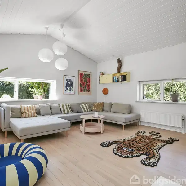 Stue med en stor, L-formet sofa omkring et lille bord, omgivet af planter og vægkunst. Rummet er lyst med hvide vægge, store vinduer og trægulv prydet med et tigerformet tæppe.