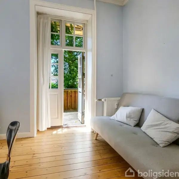 To træstole med hynder og et lille bord med en plante står på en altan. Døren til en stue står åben, og man kan se sofaen indenfor.
