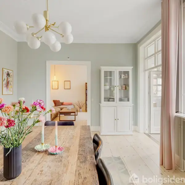 Bord med blomster i vase står i en lys spisestue med træmøbler og moderne lysekrone. Store vinduer med lyse gardiner giver et lyst miljø. En sofa ses i baggrunden.