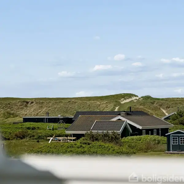 Et sort sommerhus står stille omgivet af grønne bakker og buske under en klar blå himmel med få skyer. I baggrunden ses klitter og en blå container.