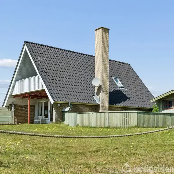 Et moderne hus står i en grøn have omkranset af et lavt hegn. Huset har et sort tag, en skorsten, og en parabol. Solen skinner på den klare himmel.