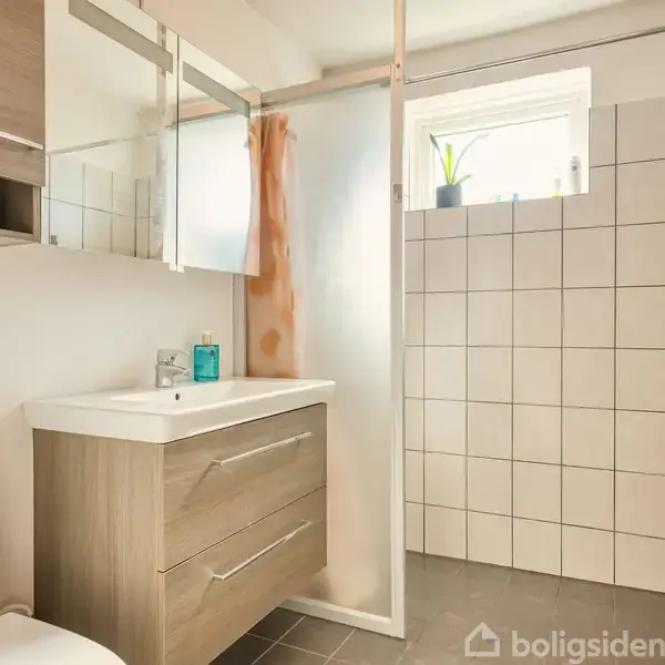 Badeværelse med hvid toilet, træskabs-vaskenhed og spejl. I brusehjørnet hænger et orange håndklæde, og vindue med plante giver lys. Flisebelagte vægge og gråt gulv.