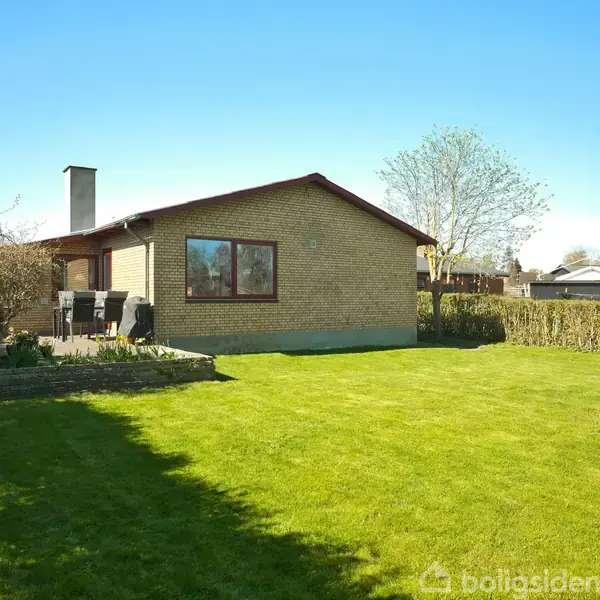 Et muret hus med skorstensrør står stille i en velplejet have med græsplæne, omgivet af hække. Der er havemøbler på en flisebelagt terrasse ved huset.