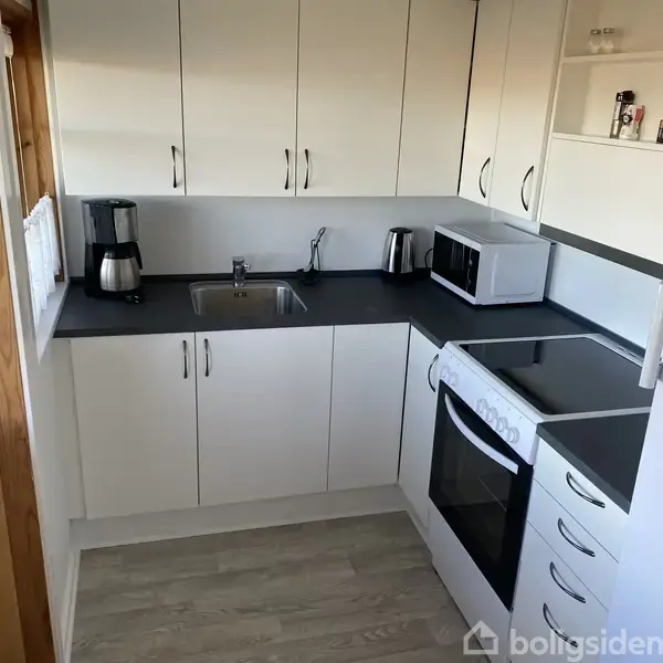 Et lille køkken med hvide skabe. En kaffemaskine, brødrister, mikroovn og komfur er placeret på sorte bordplader. Rummet er lyst med trægulv og har moderne indretning.