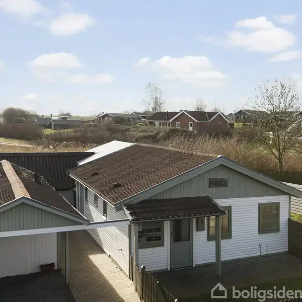 En enkeltstående hvidmalet træbygning med en carport til venstre. Beliggende i et åbent boligområde med flere huse i baggrunden under en blå himmel med spredte skyer.