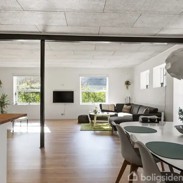 Stue med spisebord og stole, der står nær et køkkenområde. I baggrunden ses en sofa og et fjernsyn på væggen. Store vinduer lukker lys ind, der skaber en lys atmosfære.