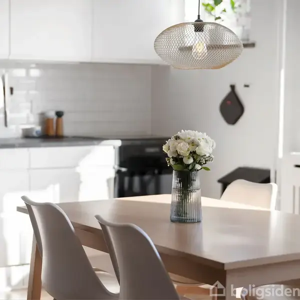 Et træbord med fire hvide stole står i et lyst køkken. En vase med hvide blomster er centreret på bordet. Over bordet hænger en moderne loftlampe.
