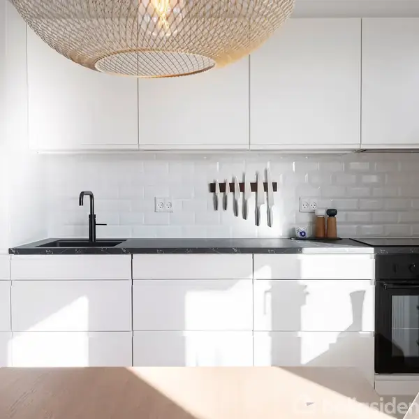 Et moderne køkken med hvide skabslåger og sort bordplade. En håndvask og en række ophængte knive ses. Rummet er oplyst af naturligt lys, og der hænger en stor rund lampe.