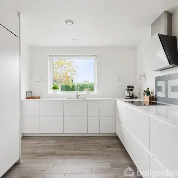 Hvidt, moderne køkken med indbyggede apparater og en l-formet arbejdsbænk. Store vinduer giver naturligt lys, mens et trægulv tilføjer varme til det rene, minimalistiske design.