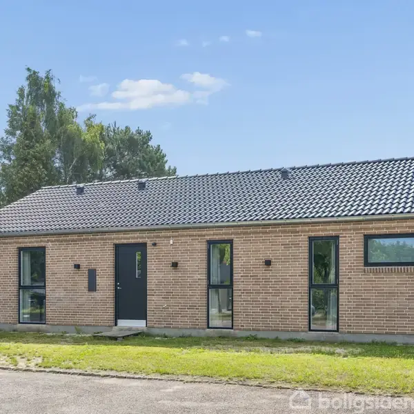 Et enkelt etplanshus med rød murstensfacade og sort tegltag står på en græsplæne. Fire store vinduer og en mørk hoveddør vender mod gaden.