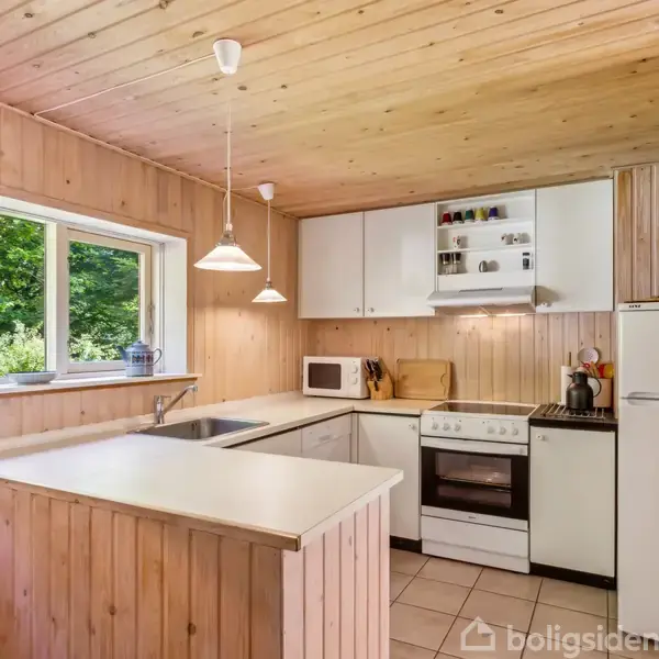 Køkken med hvidt inventar, komfur, mikroovn og køleskab. Trævægge og -loft dominerer rummet, mens vinduer lader naturligt lys ind. Ophængte lamper skaber hyggelig atmosfære. Udsigt til grønne omgivelser.