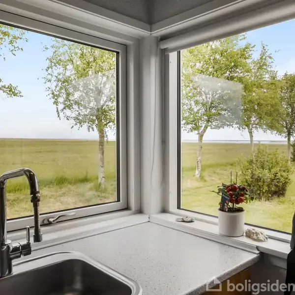 Vasken står ubrugt i køkkenhjørnet, omgivet af en lys bordplade. Udenfor ses en grøn mark, træer og en blå himmel gennem vinduerne, sammen med en kikkert og en plante på vindueskarmen.