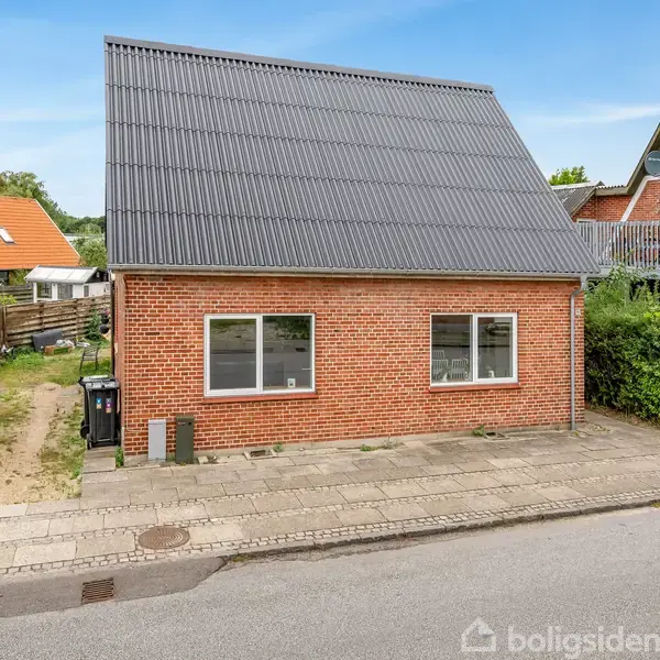 Rødt murstenshus med sort skråt tag står stille langs en fortovskant. To store vinduer vender mod gaden. Omkringliggende huse og grønt buskads i baggrunden.