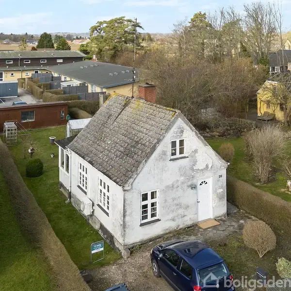 Et hvidt, gammeldags hus med gavltag står stille på en græsplæne omgivet af en lav hæk. En bil er parkeret i indkørslen foran huset.