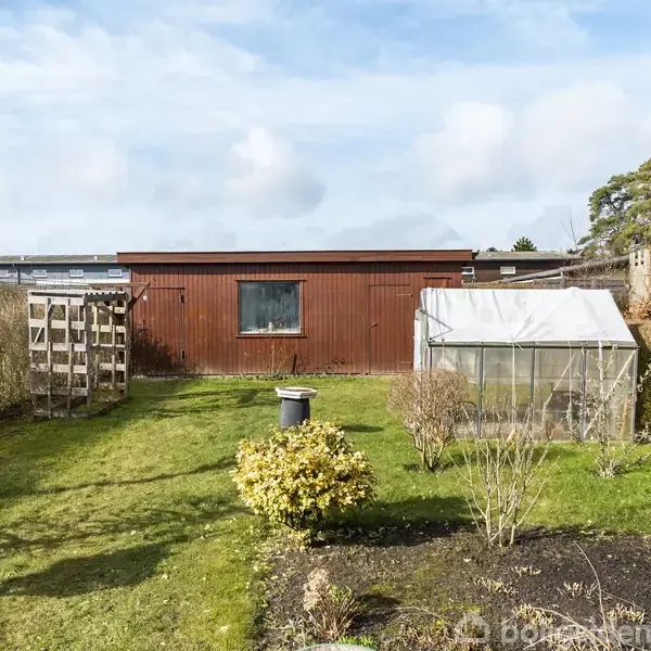 En træbeklædt bygning står midt i en velholdt have med græsplæne, drivhus og en affaldsspand. Omgivet af buske og et hegn, under en blå himmel med skyer.