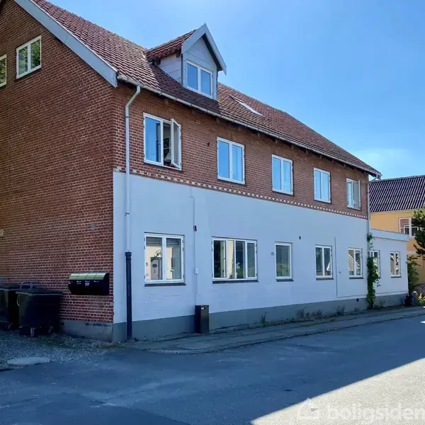 En hjørnebygning i mursten og pudsede vægge står langs en rolig gade; der er flere vinduer og et skråt tag. Ved siden af bygningen er der grøn beplantning.