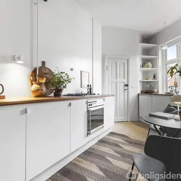 En moderne køkken med hvide skabe og træbordplade, hvor en plante og kaffemaskine står. Bord med stole ved et vindue. Udsmykning inkluderer hylder og en væglampe.