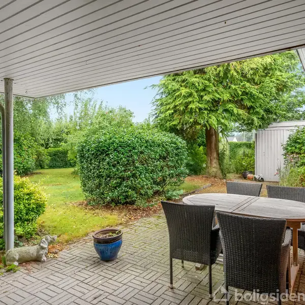 Overdækket terrasse med spisebord og seks stole. Terrasse omgivet af grøn have med buske og træer. Rødt murstenshus til højre.