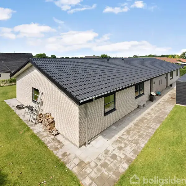 Et langt, hvidt murstenshus med sort tag står på flisebelagt areal omgivet af græsplæne og hæk under en skyfri himmel. En stige læner sig op ad husets gavl.