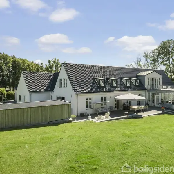 Stort hvidt hus med sort tag, liggende i en grøn have med græsplæne. Terrassen er udstyret med møbler og parasol. Omgivet af træer og blå himmel.
