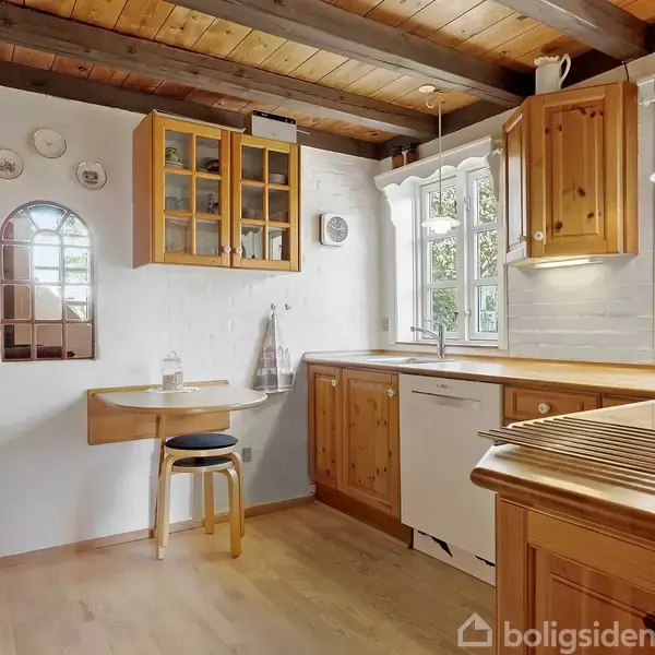 Trækøkken med træbeklædte skabe og bordplader. Et rundt bord med en stol står i hjørnet. Vinduer giver udsigt til grønne træer og omgivende natur.