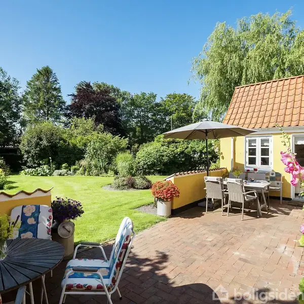 Terrassebord med stole og parasol står på en flisebelagt terrasse foran et gult hus. Foran er en velplejet have med græsplæne og blomsterbede omgivet af frodige træer.