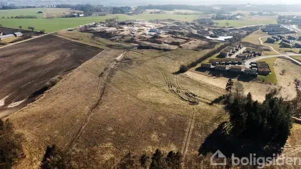 En mark ligger forladt med kørespor omgivet af træer i forgrunden. I baggrunden ses en byggeplads og et boligområde under en lys himmel og åbne marker.