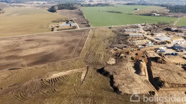 Et område under konstruktion viser jordarbejde og delvist byggede strukturer i udkanten af en by. Omkringliggende landbrugsmarker strækker sig mod horisonten under en klar himmel.