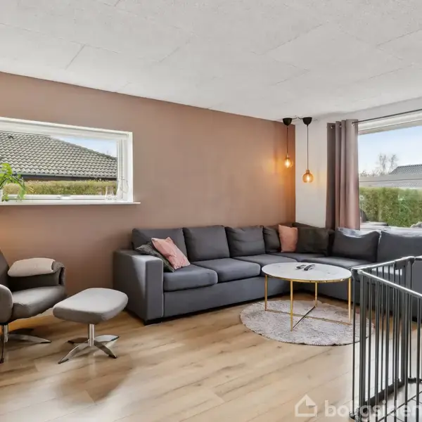Grå sofa står i hjørnet af en lys stue med trægulv, foran et sofabord. Ved siden af er en lænestol med fodskammel. Store vinduer giver udsigt til en terrasse og have.