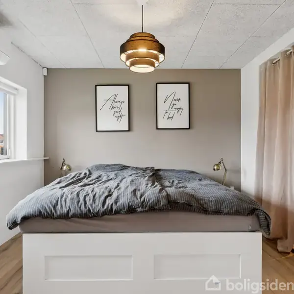 En seng placeret mod en grå væg med to lamper og to billeder, teksten: "Always kiss me goodnight" og "Never stop trying". Lys indstrømmer gennem to vinduer.