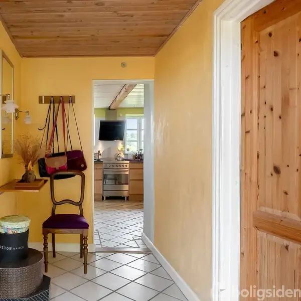En stol med en lilla hynde står i en gang med gule vægge og trægulv. En indgang fører til et køkken med et vindue og apparater. Der er ingen tekst at transskribere.
