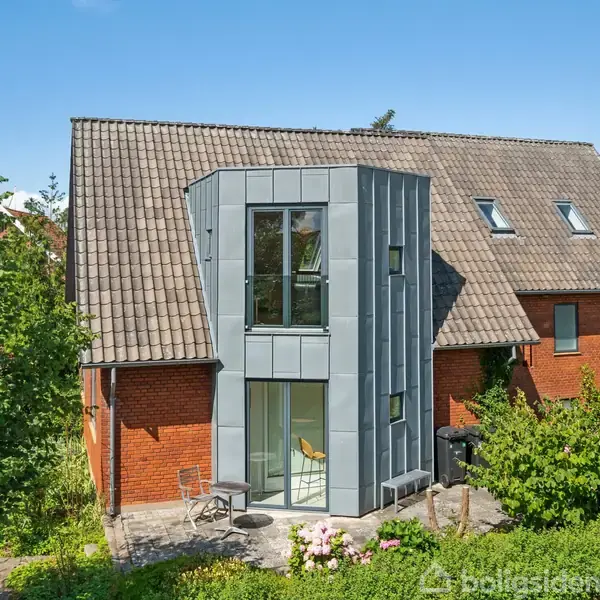 Huset er en rød murstensbygning med et moderne, gråt udbygningstårn. Tårnet har store vinduer. Huset står i en have med buske og blomster under en blå himmel.