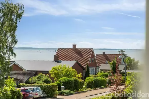 En række huse med røde tage står roligt i et grønt boligområde ved en sø eller hav, der strækker sig mod horisonten under en blå himmel med få skyer.