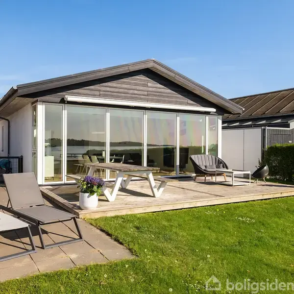 Et moderne sommerhus med store glaspartier står stille i en solrig have. To liggestole og et bord-bænkesæt er placeret på en træterrasse omgivet af velplejet græs og buske.
