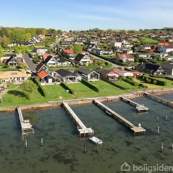Kystnære huse står i række langs en bred græsplæne ved en stille havn med bådebroer. Bag husene strækker en grøn by med mange boliger sig op ad en bakke.