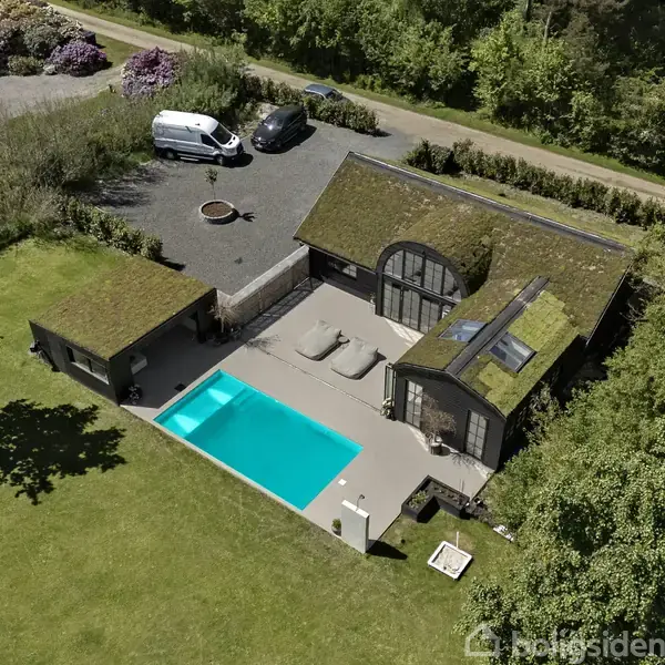 Hus med græs på taget står ved siden af en swimmingpool. Omkringliggende parkering har biler, inklusive en hvid varevogn. Området er omgivet af grøn frodig vegetation.