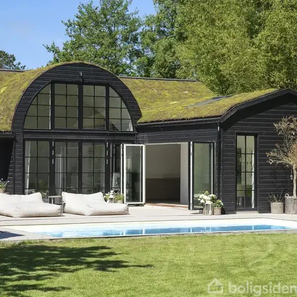 Et moderne sortmalet hus med store vinduer åbner sig mod en terrasse. To hvide liggestole står ved en swimmingpool, omgivet af grønne træer og græsplæne.