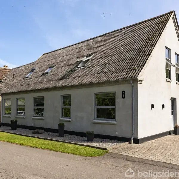 Et hvidt, vinkelformet hus med skråt tag og flere ovenlysvinduer. Huset står ved en vej med grønne buske og klar blå himmel i baggrunden. Nummer "6" er synligt på væggen.