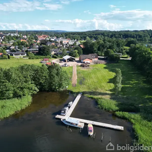 En bådebro strækker sig ud i en sø med flere både fortøjet. Omgivet af grønne marker og træer i nærheden af en by med huse og bygninger i baggrunden.
