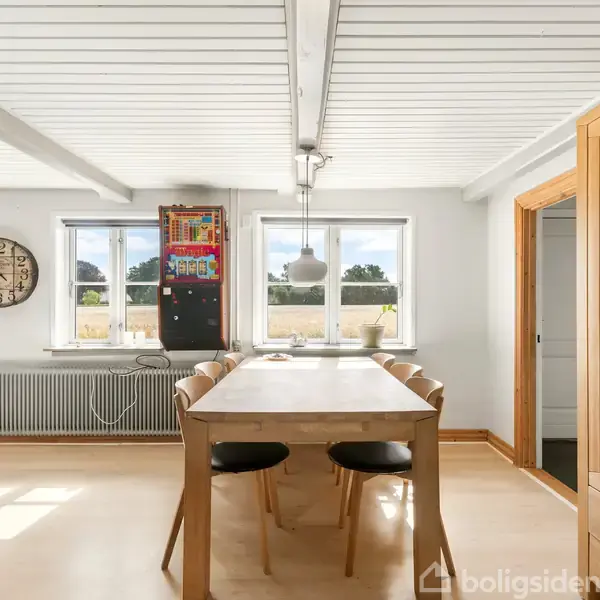 Et rektangulært spisebord med seks stole står i et lyst rum med trægulv. Vinduerne bagved viser landskab udenfor. Væggen har et stort ur og farverigt vægpanel.