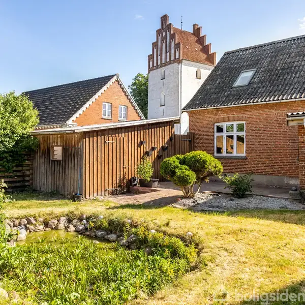 Murstensbygning med trædør i midten, omgivet af to store vinduer på hver side. En postkasse er monteret på væggen. Bygningen har et mørkt tegltag, og fortovet er synligt foran huset.