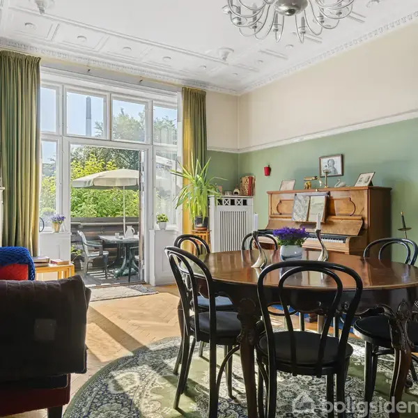 Rundt spisebord med sorte stole står på tæppe i stue. Et klaver står ved siden af i hjørnet. Store vinduer fører ud til terrasse i sollys.
