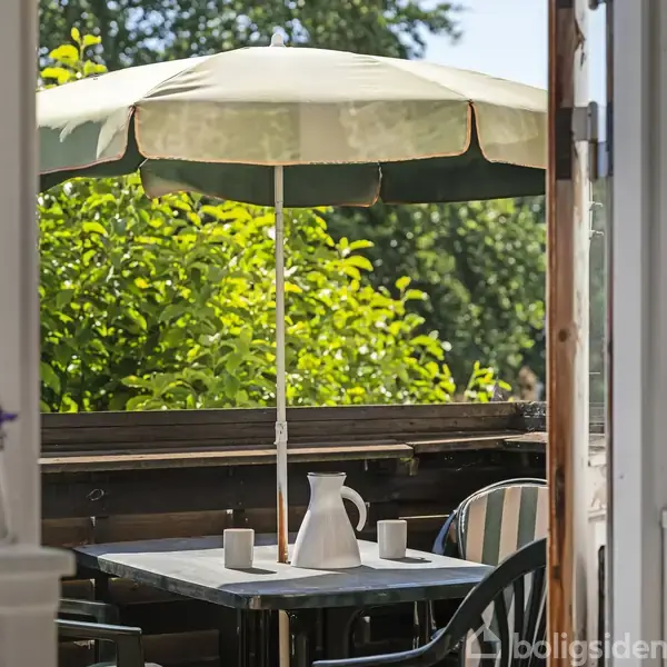 En terrasse har et bord med to stole dækket af en hvid parasol. En kande og krus står på bordet. Omkring er der grønne buske og planter.