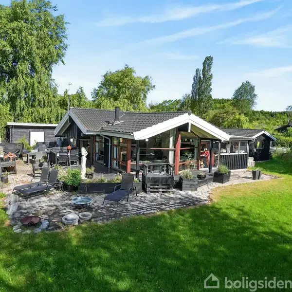 Hus med mørkt tag og lyse paneler står i en frodig have med stor terrasse. Terrasse har flere havemøbler. Omkringliggende træer giver en naturskøn setting.
