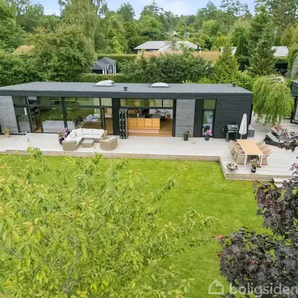 En moderne sort-hus med store vinduer står på en træterrasse med havemøbler. Græsplæne og tæt bevoksning omgiver huset i en hyggelig, grøn have.
