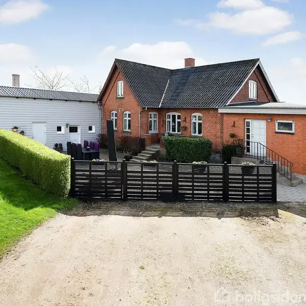 Rødt murstenshus med sort tag står stille omgivet af grønne græsplæner. En sort stakitport foran huset markerer indgangen. Hvid bygning ved siden af hovedhuset.